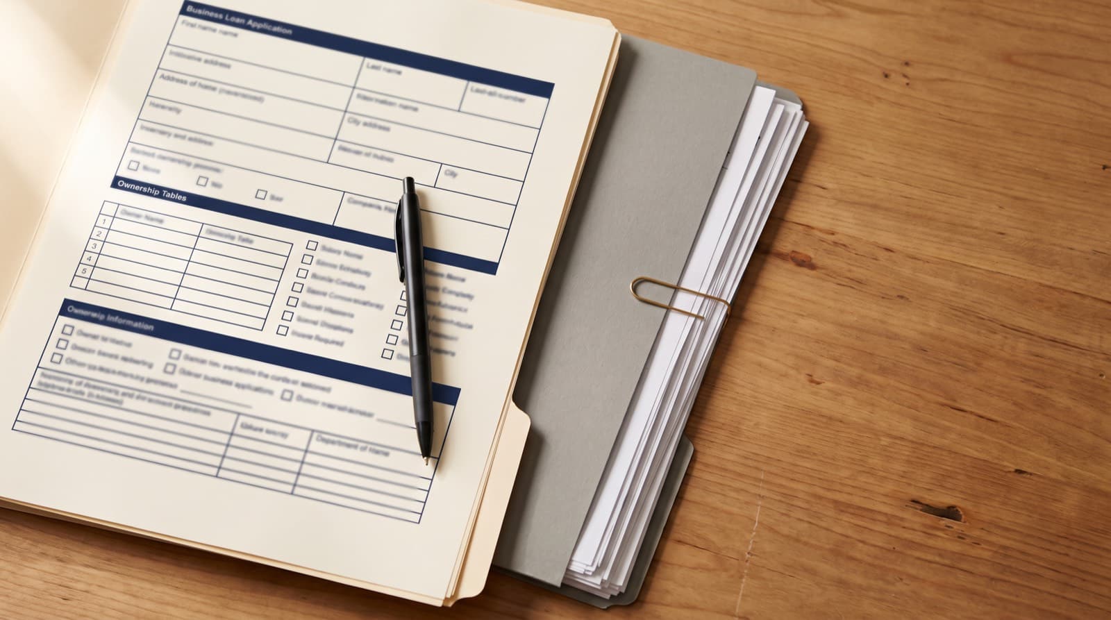 A business loan application form with an ownership table and eligibility checkboxes, a pen resting on the page beside a folder