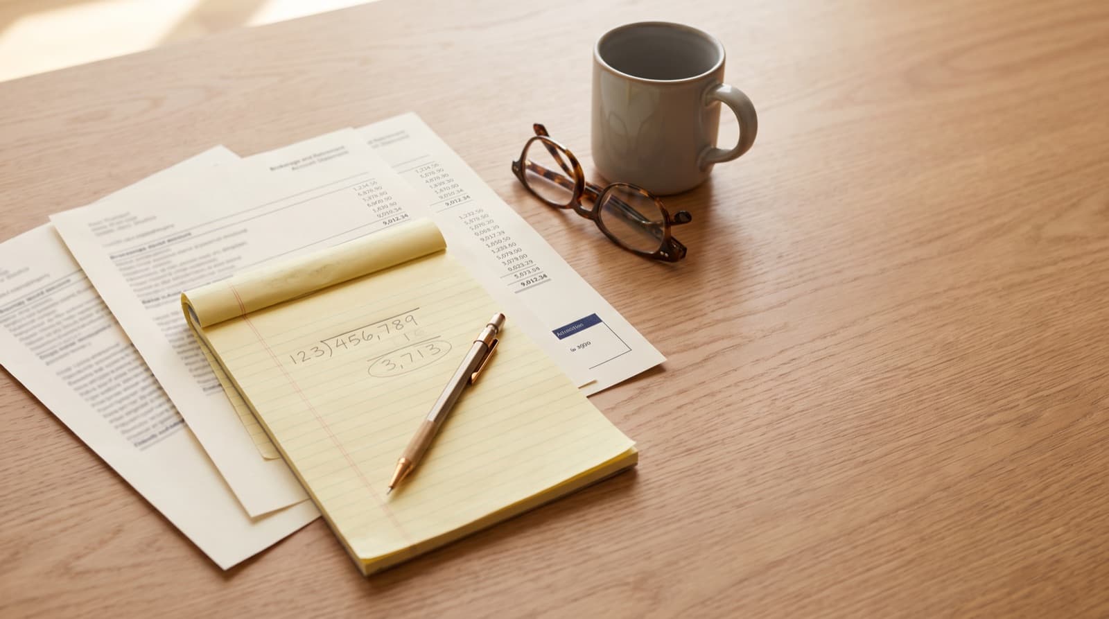 Brokerage and retirement account statements fanned across a wooden desk beneath a yellow legal pad showing a handwritten long-division calculation, next to reading glasses and a coffee mug