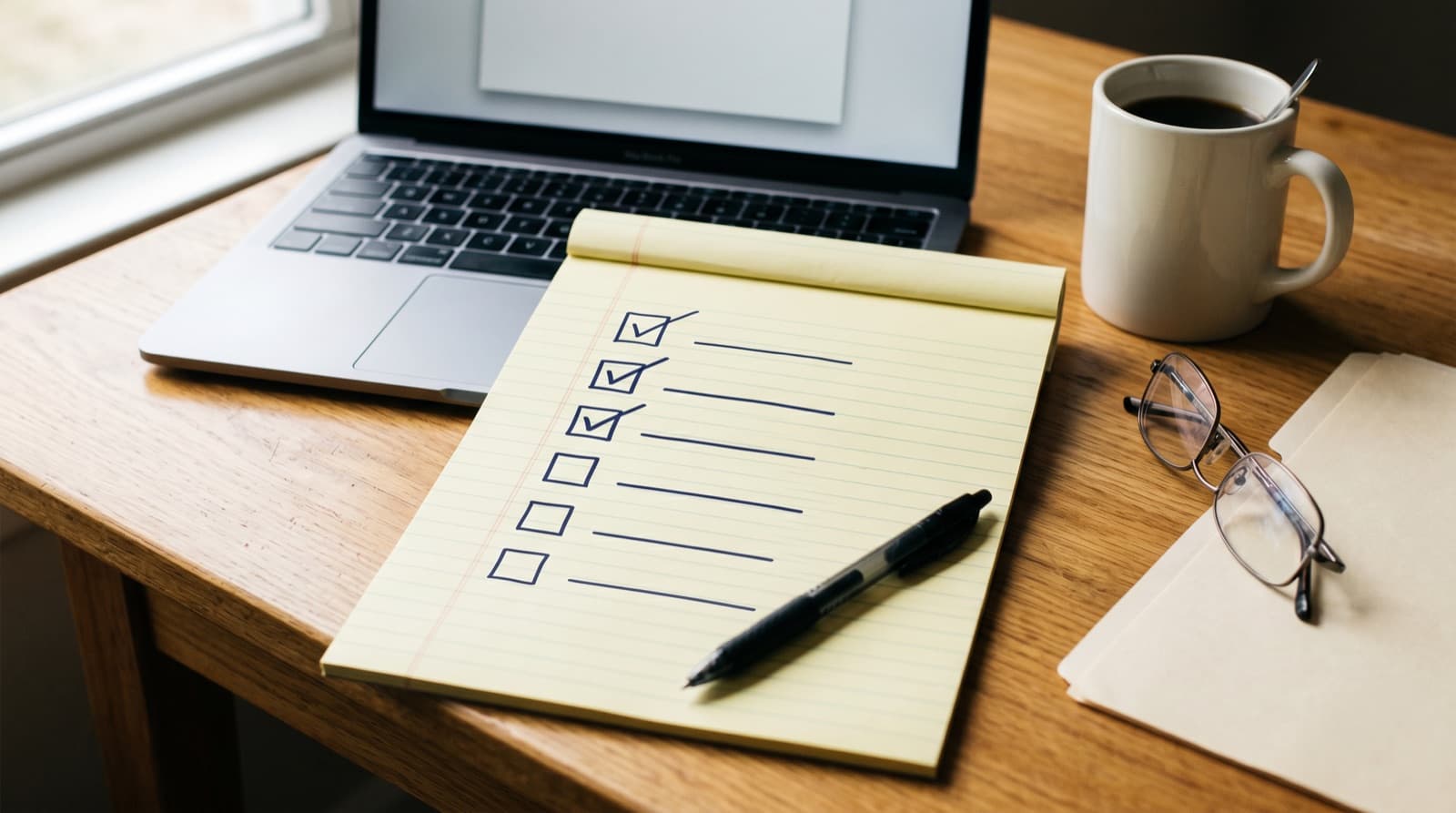 A yellow legal pad lying on a warm wooden desk beside an open laptop, with a six-item checkbox list drawn in navy ink, the first three boxes checked